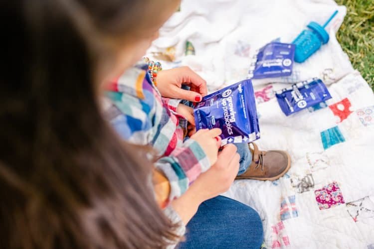A young girl holds prebiotic sachets outdoors on a quilt.