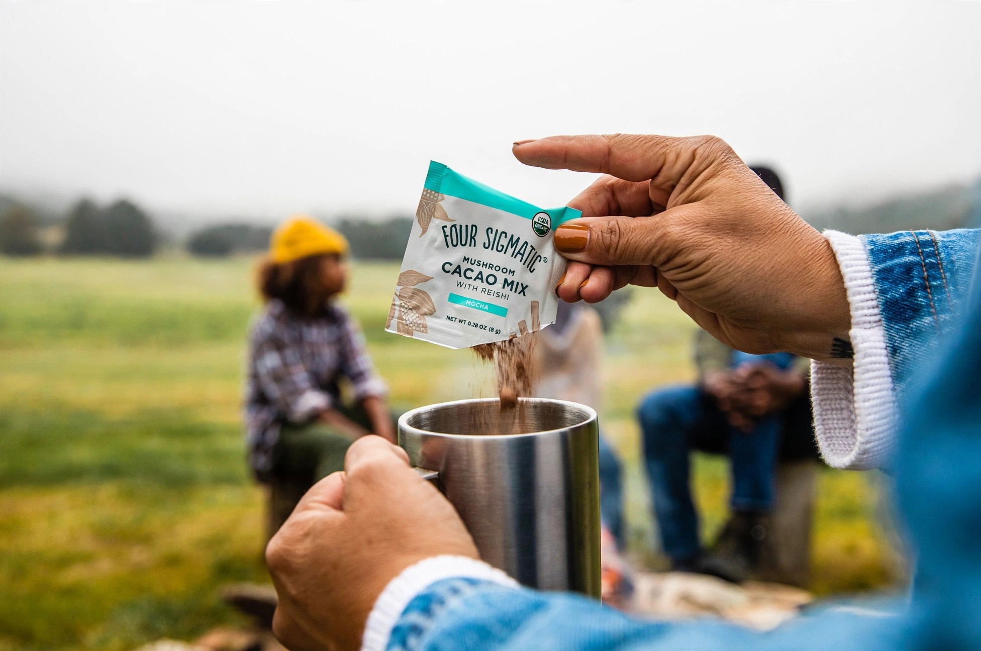 A person empties a cacao packet into a metal cup outdoors.