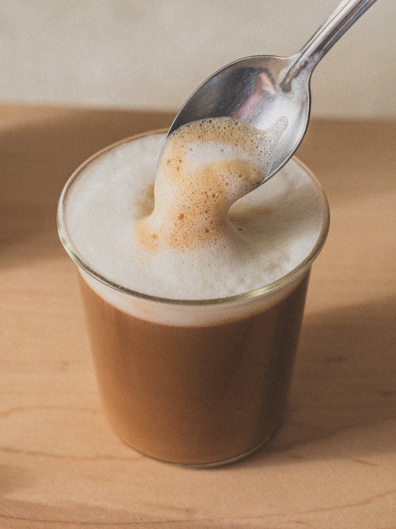 Spoon holding frothed milk over a small glass latte on table.